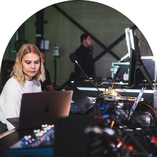 Achter de schermen van een live mediaproductie met jonge vrouwelijke technicus die werkt op een Apple MacBook in een controle- of regieruimte. De vrouw met halflang blond haar en witte trui kijkt geconcentreerd naar haar laptopscherm terwijl ze omringd is door audiovisuele apparatuur, kabels, een mengpaneel en netwerkverbindingen. Op de achtergrond staan collega’s en professionele studioapparatuur met monitoren en microfoon. Sfeer van eventproductie, technische ondersteuning, livestream-setup, broadcast control room, IT- en AV-technologie in een industriële binnenlocatie.